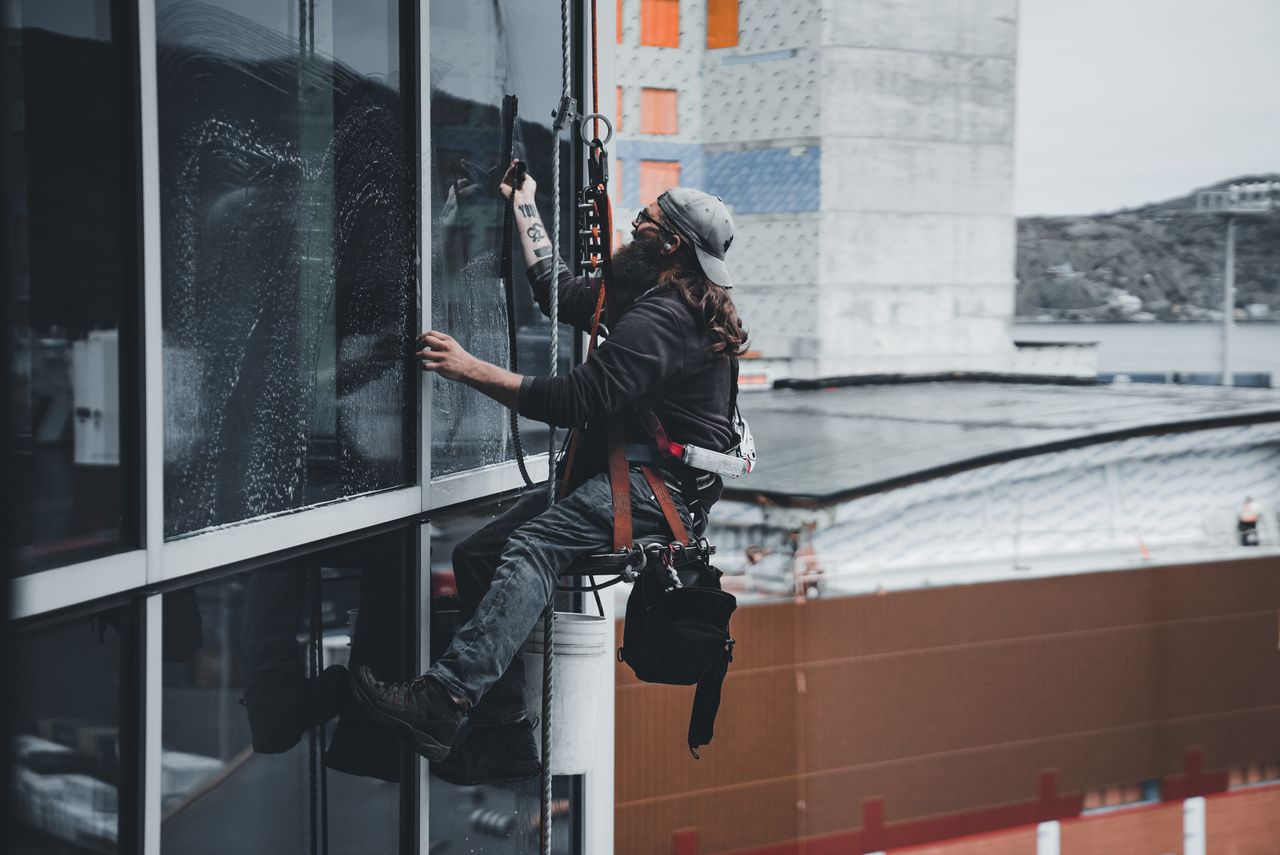 gallery-03 A man cleaning skyscraper windows with a harness, showcasing urban high-rise work.
